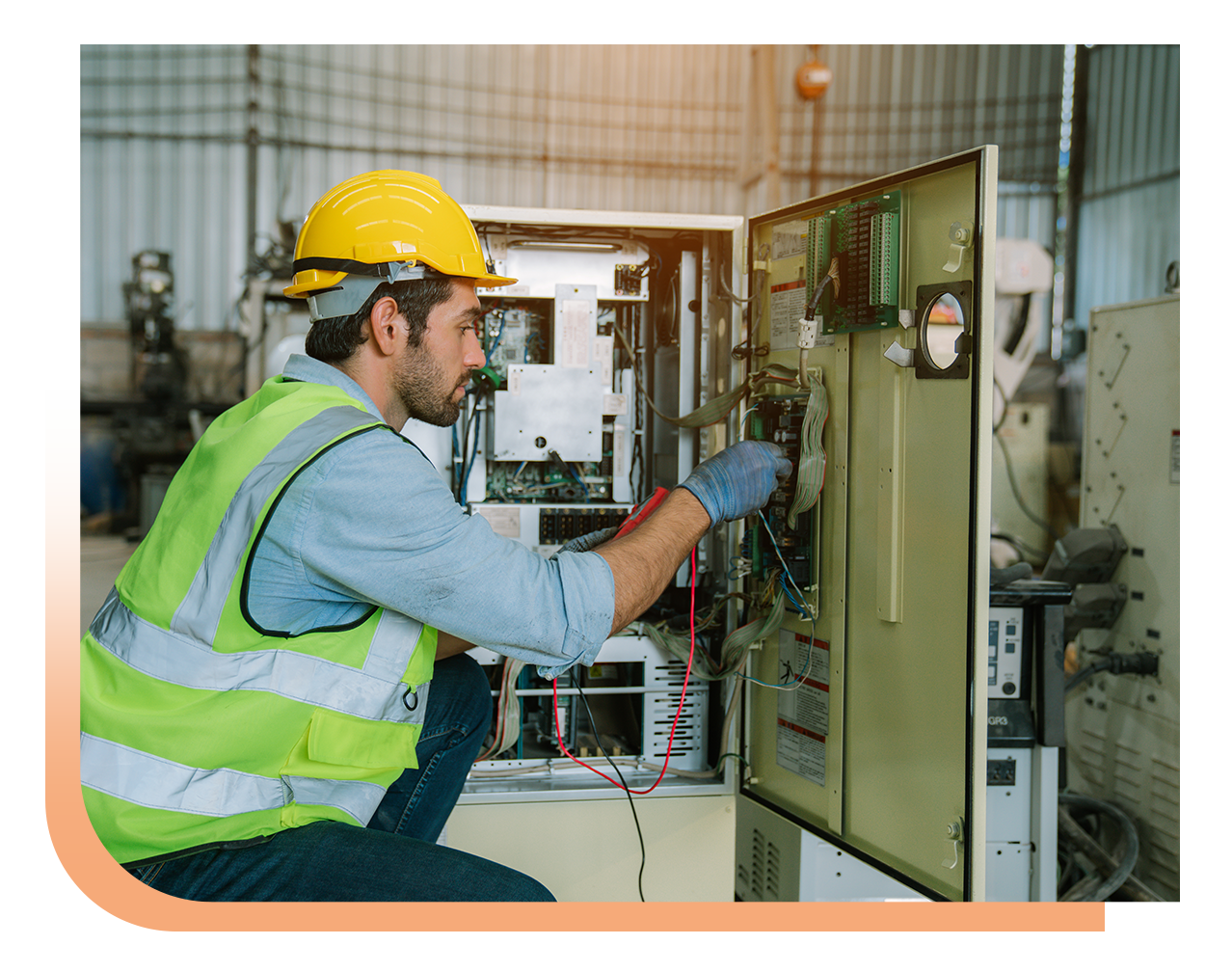 Technician in a yellow hard hat and safety vest working on an open electrical control panel in a factory setting.