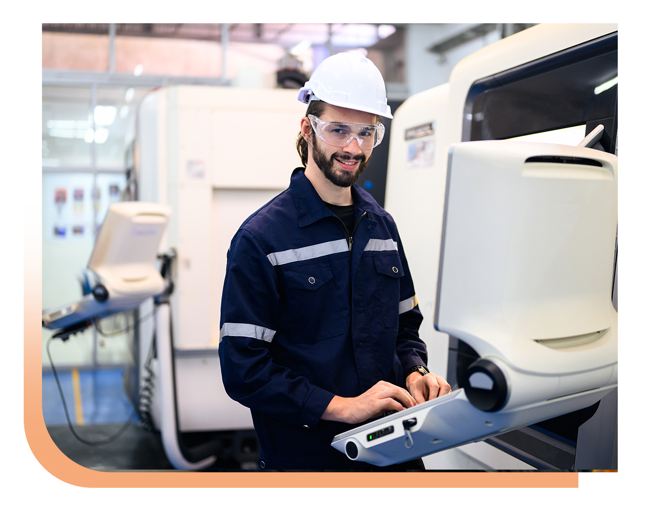 Male technician in a hard hat and safety glasses operates a control panel beside a large industrial machine in a factory setting.
