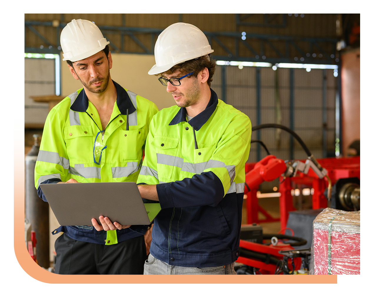 Two construction workers in neon safety jackets and white hard hats review a laptop together in a workshop.