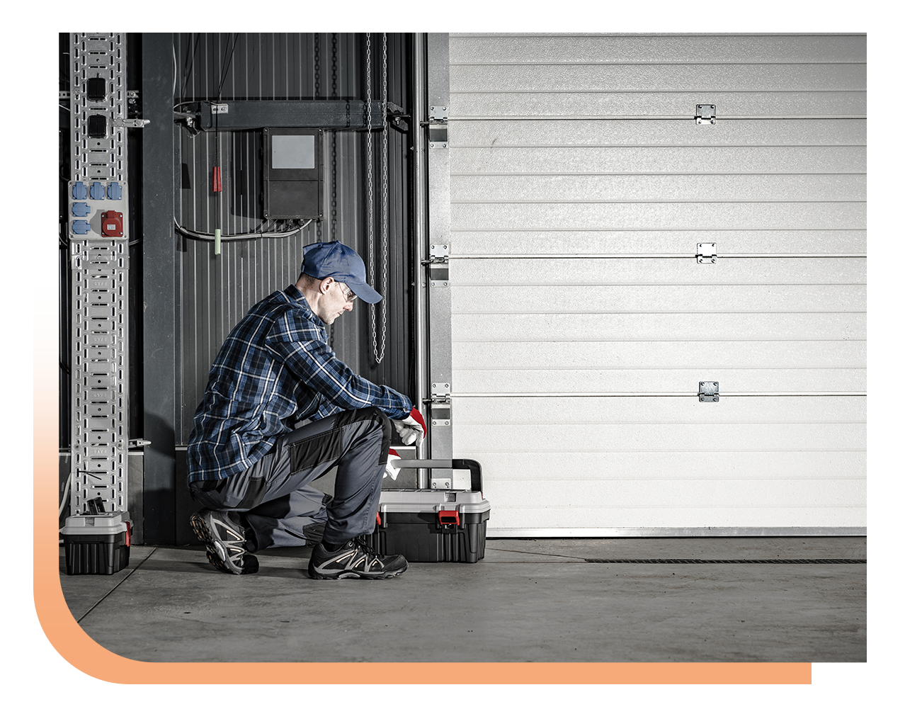 Maintenance technician kneeling by a large garage door, using a toolkit for repairs beside a toolbox.