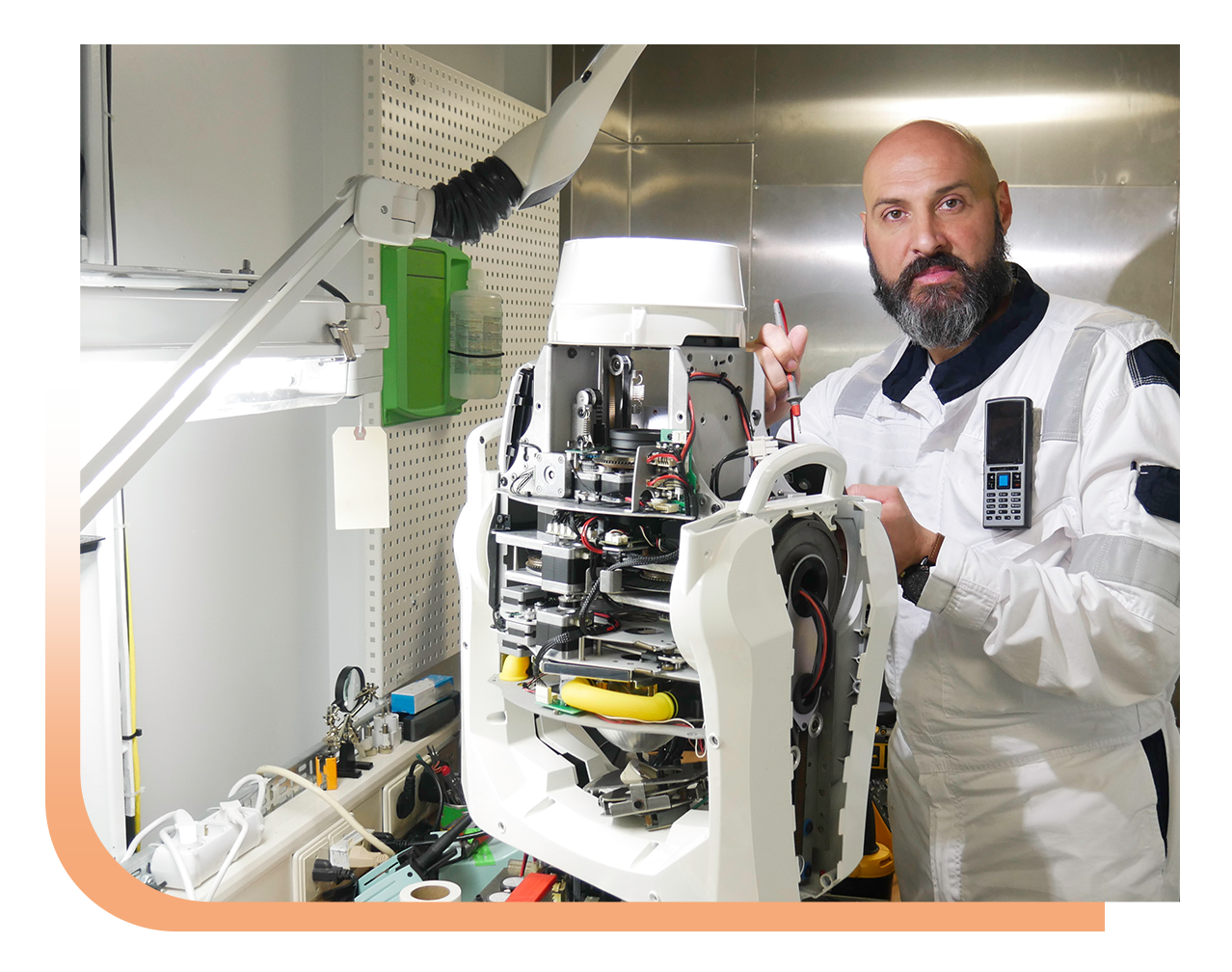 Bearded technician in white coveralls repairing a disassembled white robotic device on a workbench in a lab.