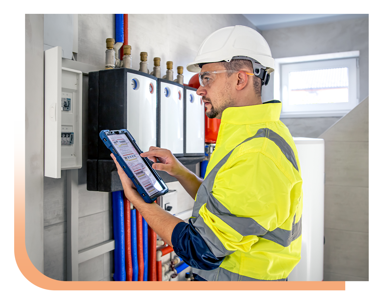 Male technician in a hard hat and high-visibility jacket using a tablet at an electrical control panel.