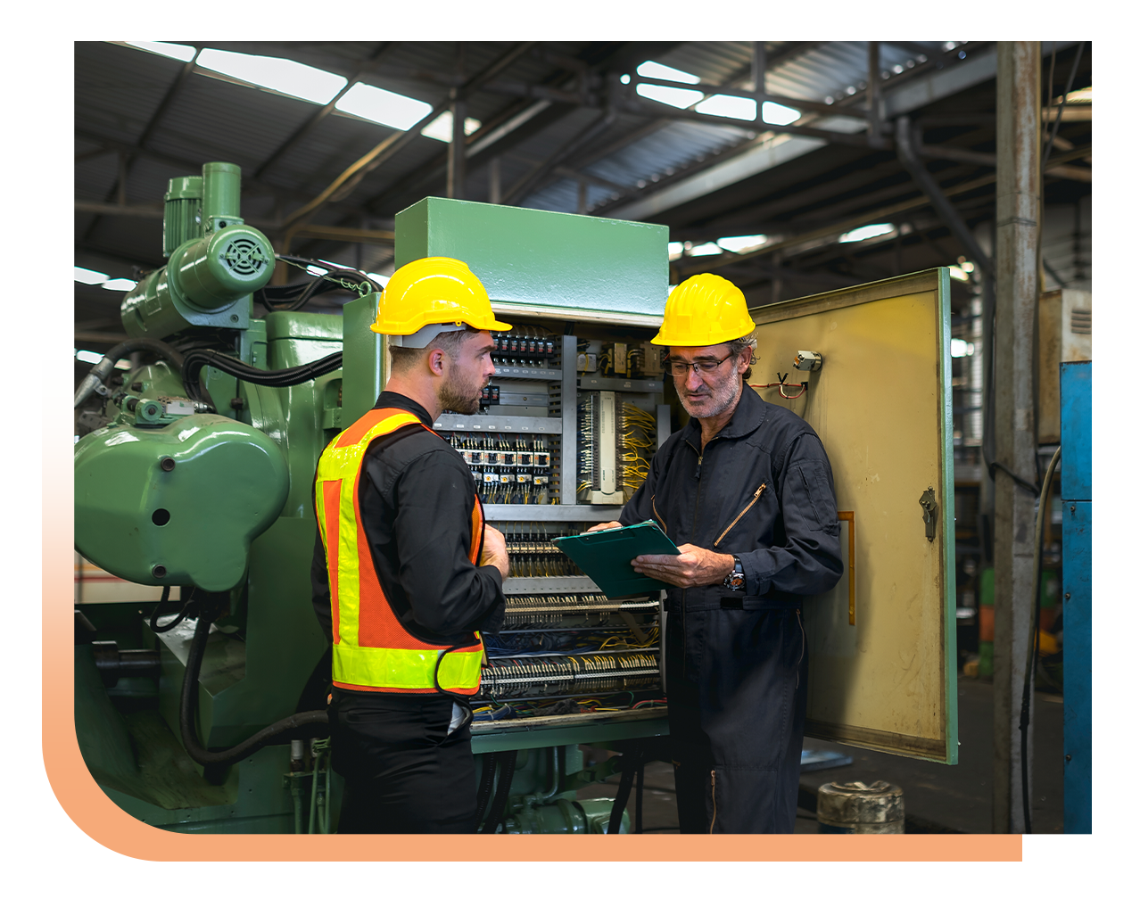 Two workers with yellow hard hats inspect an open control panel in a factory, one holding a clipboard and discussing with the other.
