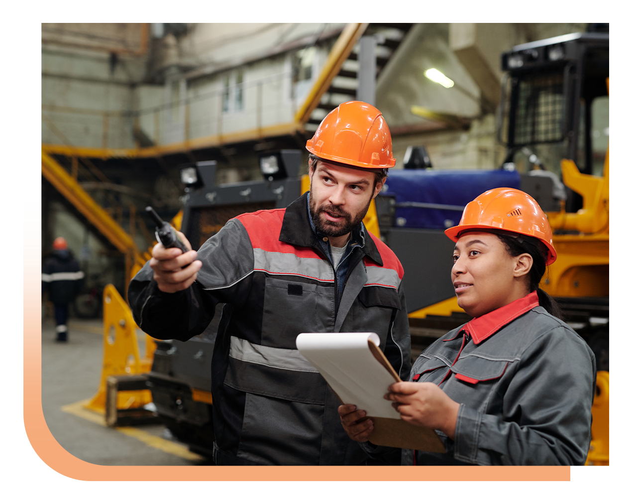 Two factory workers in orange hard hats review plans; one points with a tool while the other holds a clipboard in a busy industrial workspace.