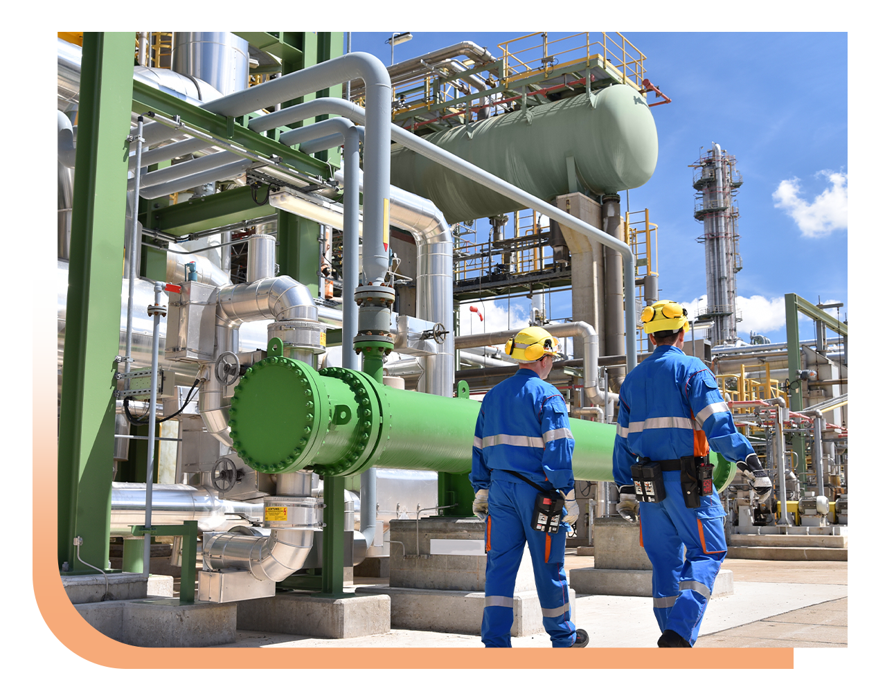 Two workers in blue coveralls and yellow hard hats walk beside a large green industrial valve and network of silver pipes at an outdoor plant.