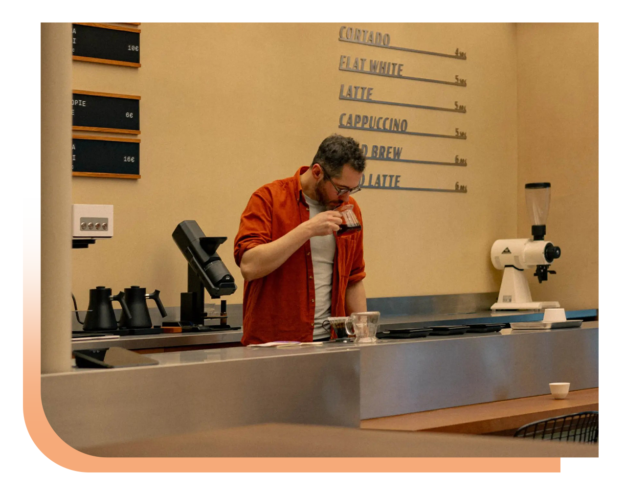 Barista in an orange shirt tasting coffee at a cafe counter, with a grinder and espresso machine visible in the background.