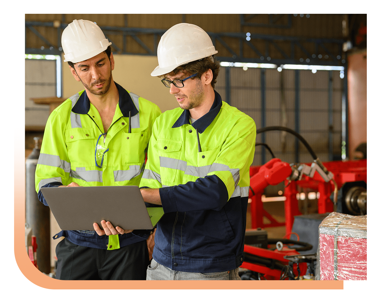 Two construction workers in neon safety jackets and hard hats review information on a laptop in an industrial workshop.